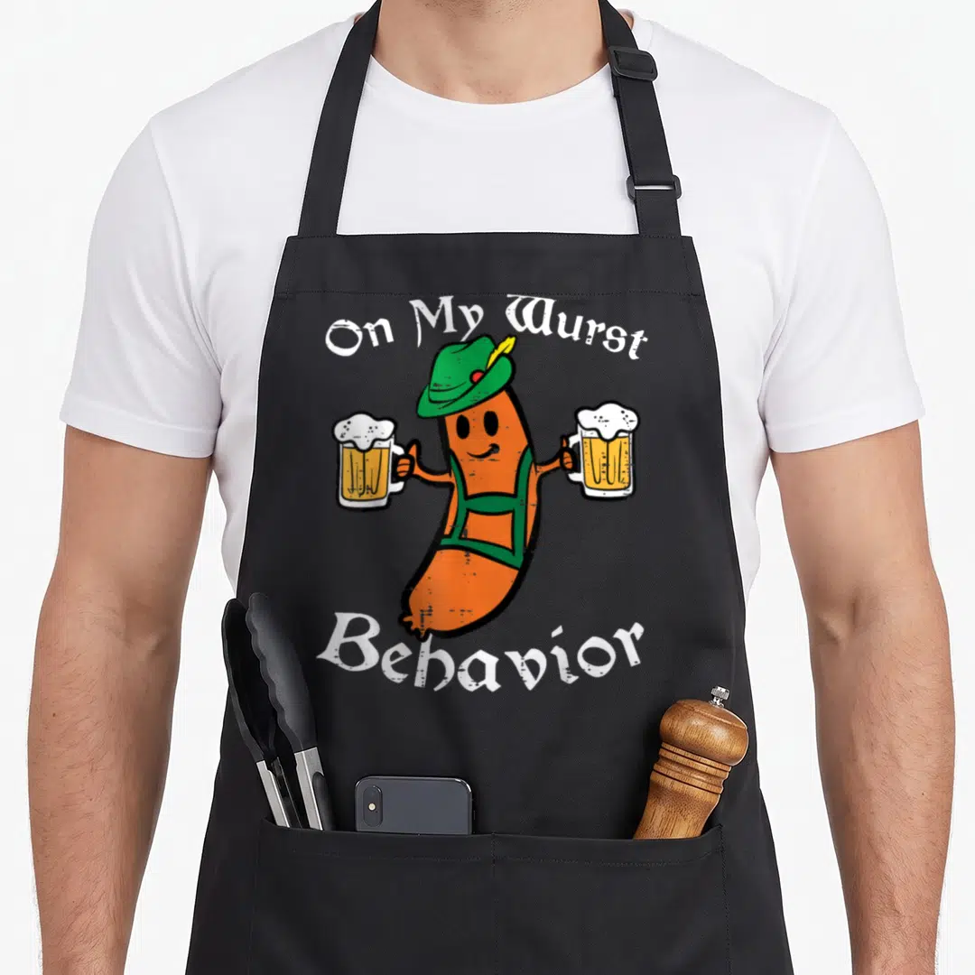 Wurst german apron - Cartoon orange sausage in green hat holding two beer mugs with white text On My Wurst Behavior — shown on a black cotton canvas apron against a white background