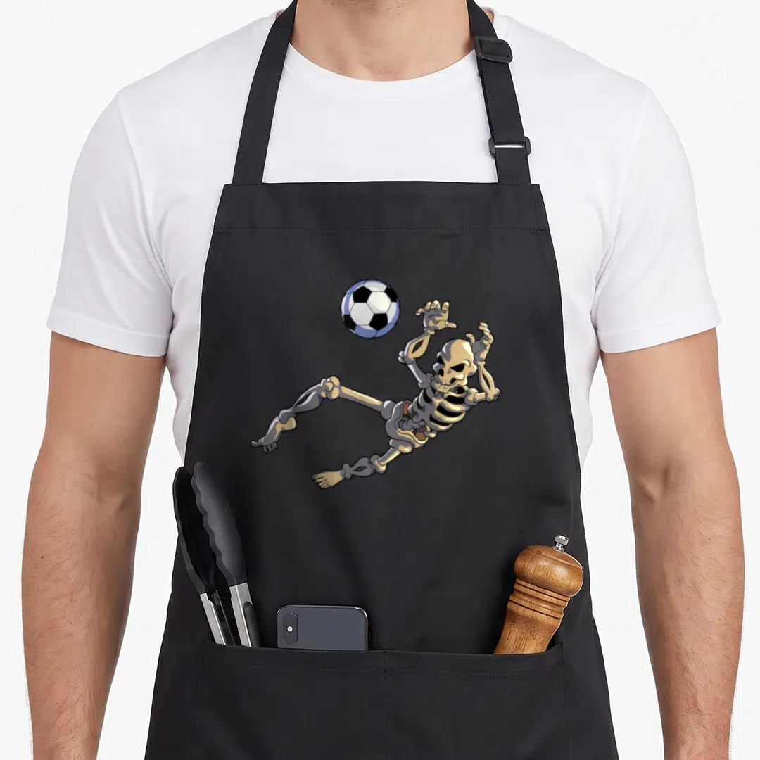 Goalkeeper halloween apron - Cartoon skeleton reaching for soccer ball in beige white and black — shown on a black cotton canvas apron against a white background