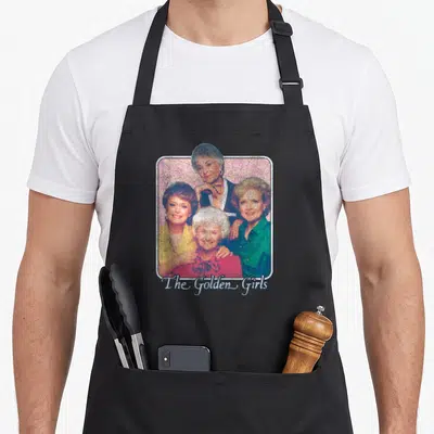 Iconic golden apron - Four women clustered in rounded frame with pink, yellow, red, green, blue hues and centered text — shown on a black cotton canvas apron against a white background
