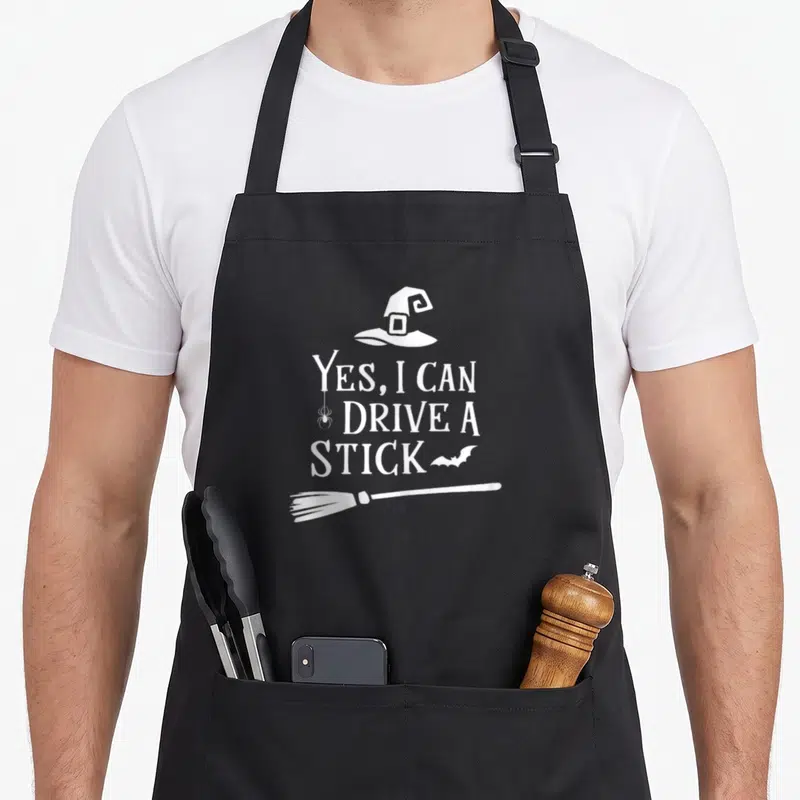Drive i love witch apron - White witch hat spider bat broomstick text Yes I Can Drive A Stick cartoon design — shown on a black cotton canvas apron against a white background