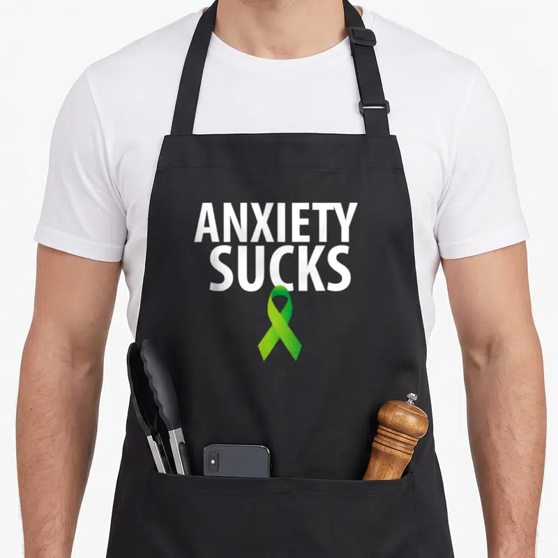Anxiety ribbon apron - ANXIETY SUCKS text above green awareness ribbon — shown on a black cotton canvas apron against a white background