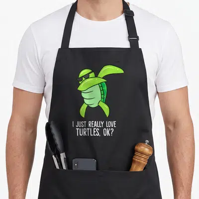 Dabbing novelty apron - Green cartoon turtle dabbing with sunglasses above white text I just really love turtles ok — shown on a black cotton canvas apron against a white background