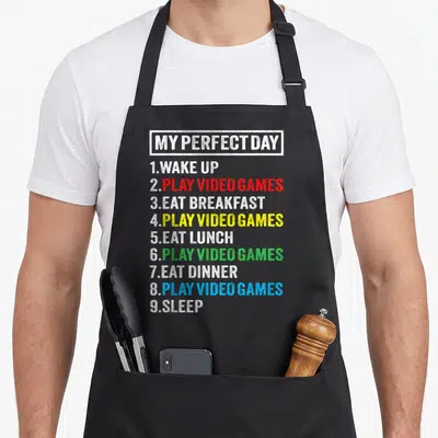 Cute perfect video game apron - Stacked text list reading My Perfect Day with Play Video Games in red yellow green — shown on a black cotton canvas apron against a white background