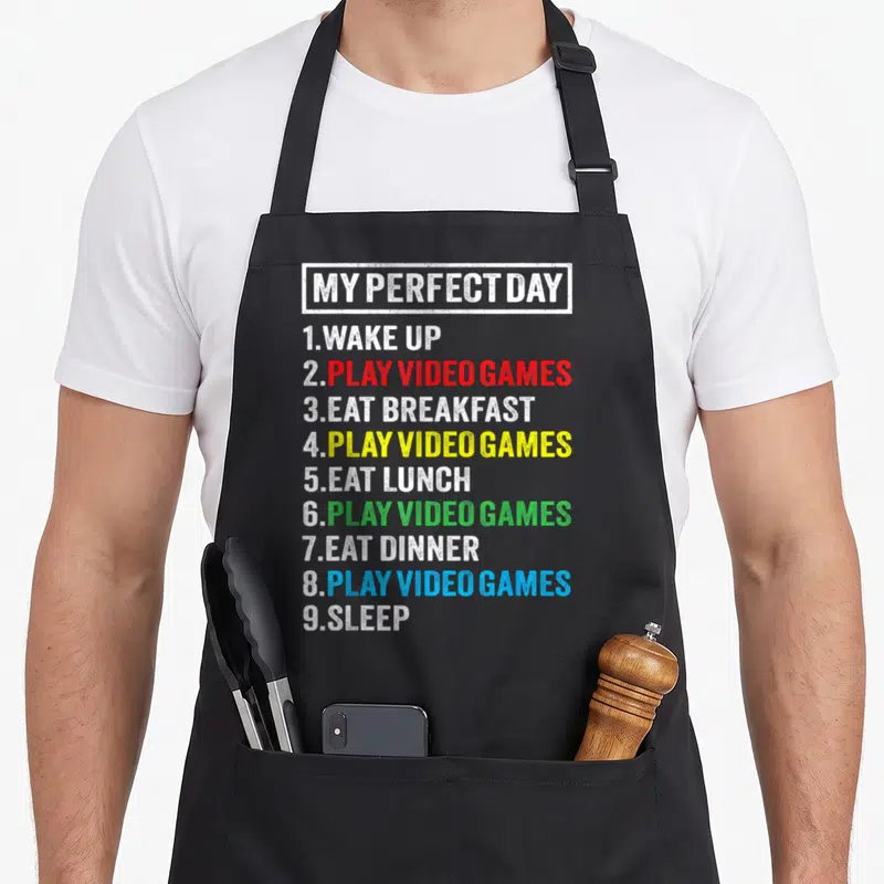 Cute perfect video game apron - Stacked text list reading My Perfect Day with Play Video Games in red yellow green — shown on a black cotton canvas apron against a white background