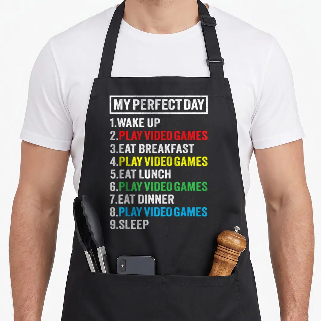 Cute perfect video game apron - Stacked text list reading My Perfect Day with Play Video Games in red yellow green — shown on a black cotton canvas apron against a white background