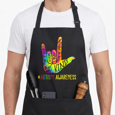 Asl tie-dye apron - Rainbow tie-dye ASL hand sign with Be Kind text and autism awareness hashtag — shown on a black cotton canvas apron against a white background