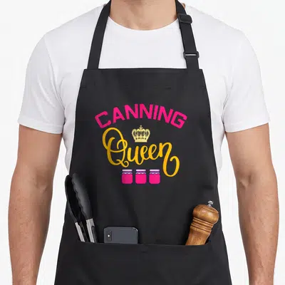 Queen canning apron - Pink arched CANNING text, yellow Queen script, gold crown, three purple jars — shown on a black cotton canvas apron against a white background