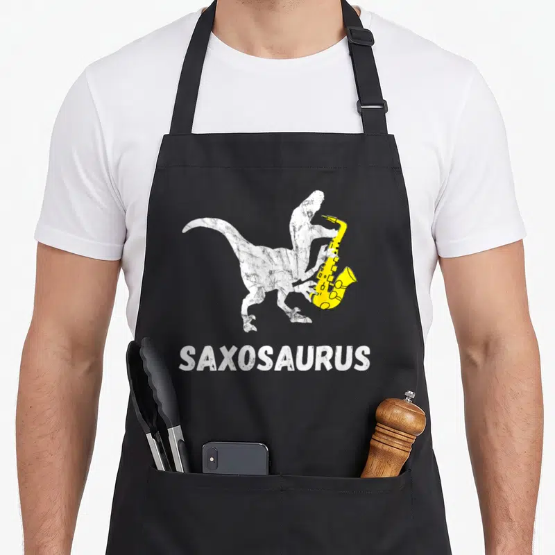 Sax dinosaur apron - White dinosaur holding yellow saxophone with SAXOSAURUS text below — shown on a black cotton canvas apron against a white background