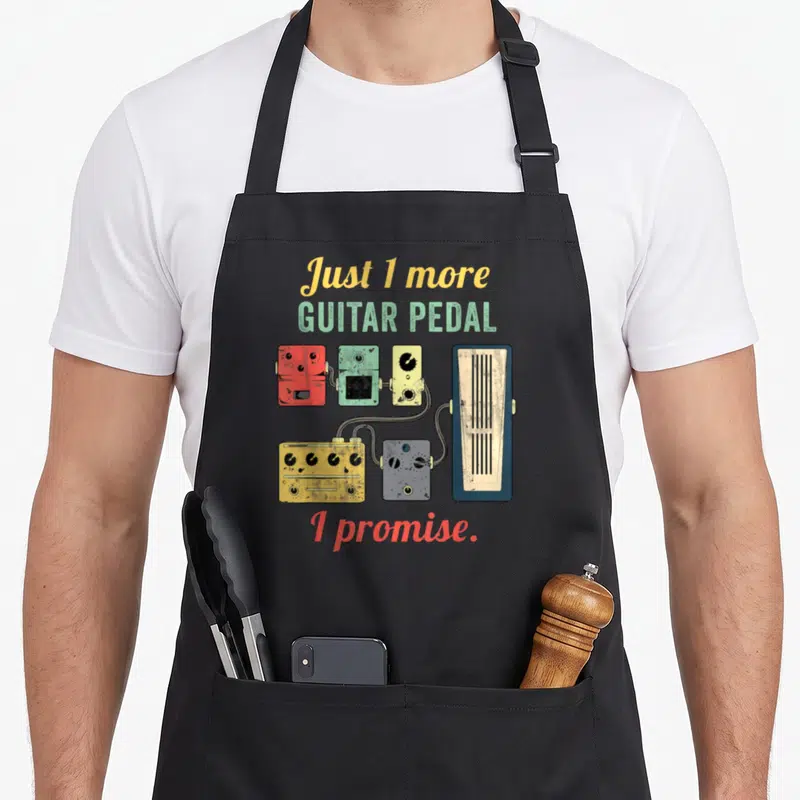 Amplifier crew apron - Cartoon guitar pedals in yellow green red and blue connected by cables under the text Just 1 — shown on a black cotton canvas apron against a white background