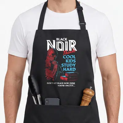 Noir superhero apron - Black Noir character in red and blue with stacked books and study quote text — shown on a black cotton canvas apron against a white background