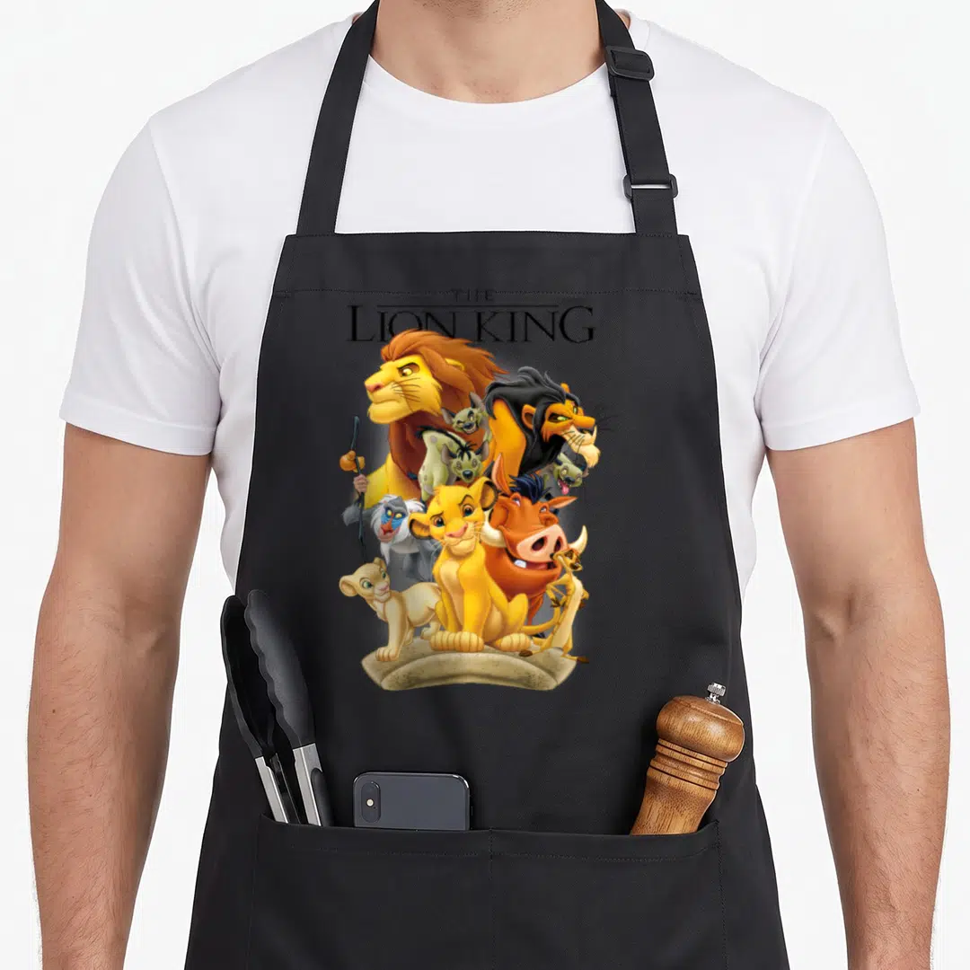 Land pride apron - Cartoon lions and text The Lion King clustered on a rock base — shown on a black cotton canvas apron against a white background