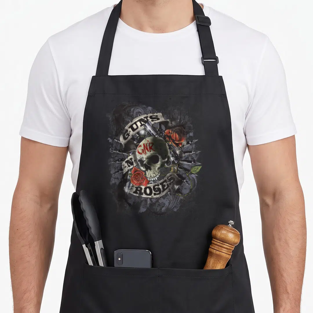 Firepower gun apron - Distressed skull with GNR text, revolver cylinder, red roses, and GUNS N' ROSES ribbon in black — shown on a black cotton canvas apron against a white background