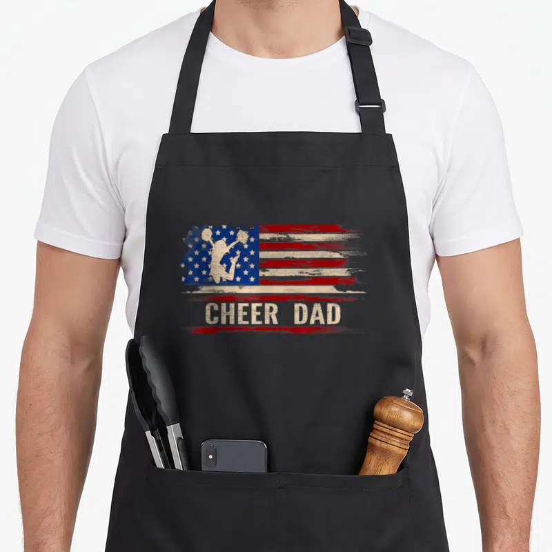 Cheer american flag apron - Distressed USA flag with cheerleader silhouette and CHEER DAD text on red brushstroke — shown on a black cotton canvas apron against a white background