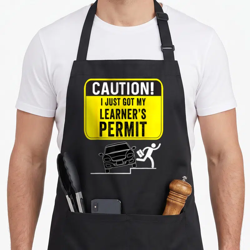 Caution student aprons - Yellow caution sign with learner permit text above a black car chasing a running figure — shown on a black cotton canvas apron against a white background