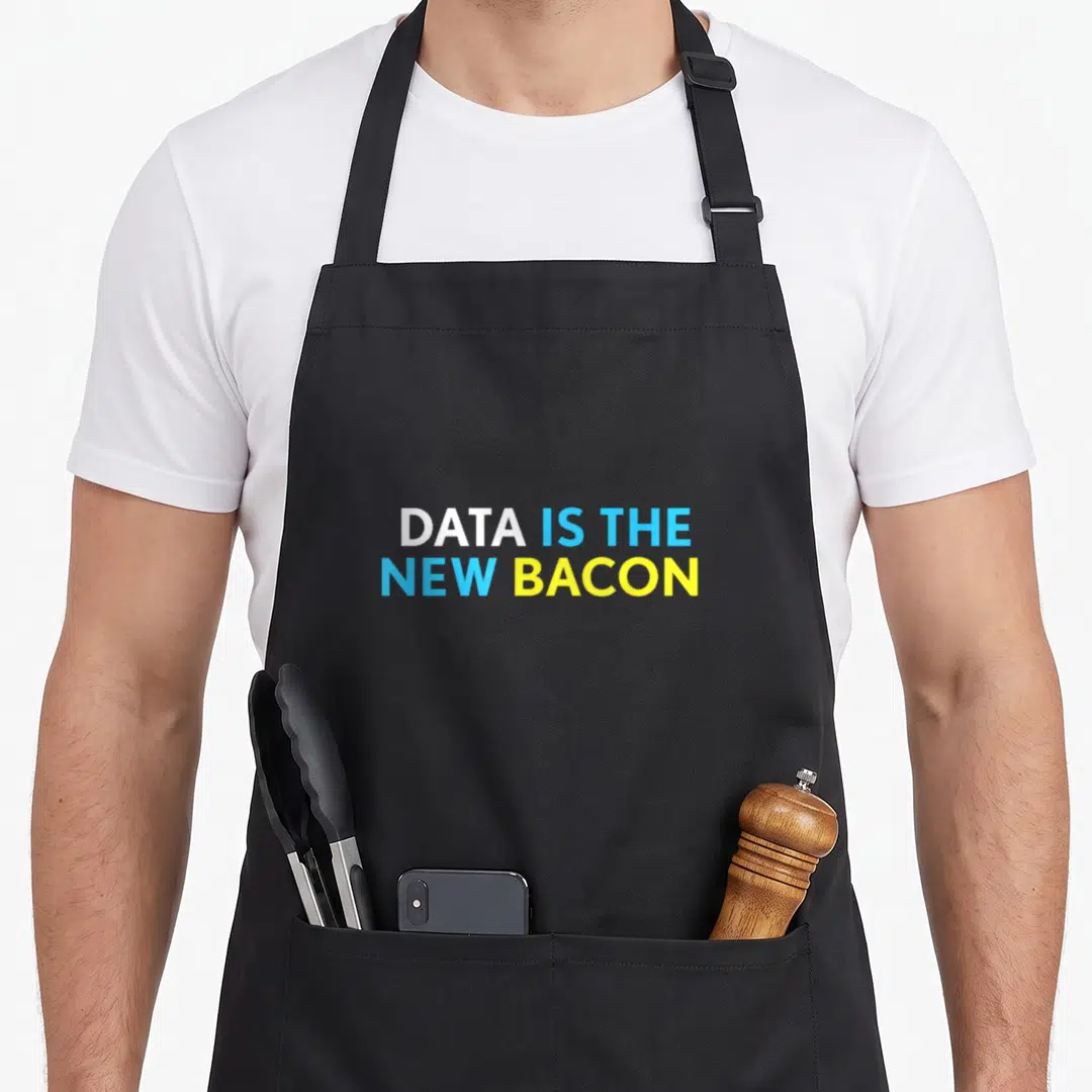 Data bacon apron - Stacked DATA IS THE NEW BACON text in white, light blue, and yellow block letters — shown on a black cotton canvas apron against a white background
