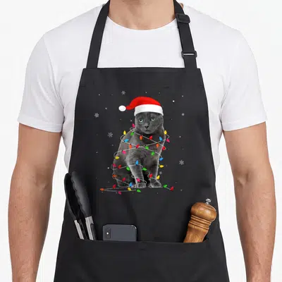 Christmas russian aprons - Grey cat wearing red santa hat wrapped in multicolored Christmas lights with white snowflakes — shown on a black cotton canvas apron against a white background