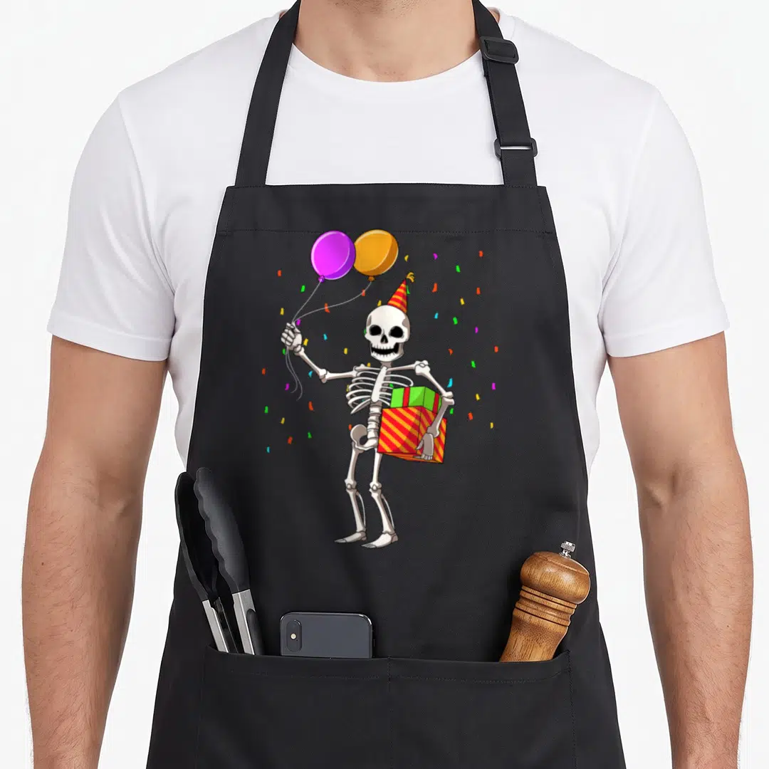 Outfit funny halloween apron - Cartoon skeleton holding purple balloon and two stacked gift boxes surrounded — shown on a black cotton canvas apron against a white background