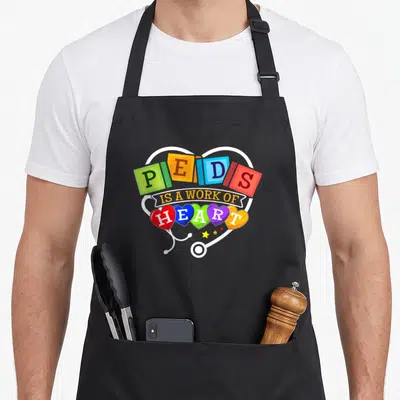 Pediatric pediatrician nurse apron - Colorful alphabet blocks and hearts spelling Peds Is a Work of Heart inside white — shown on a black cotton canvas apron against a white background