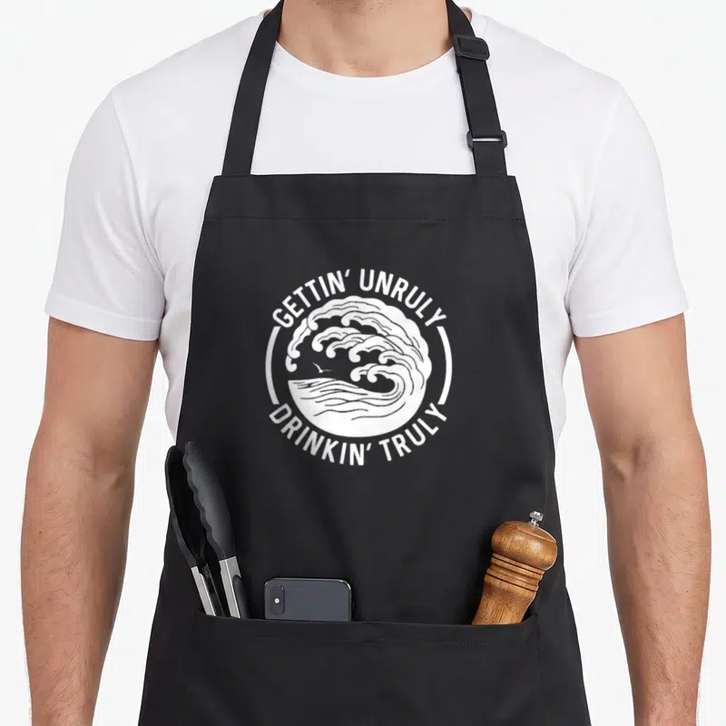 Gettin summer aprons - White circular wave design with bird and text GETTIN UNRULY DRINKIN TRULY — shown on a black cotton canvas apron against a white background