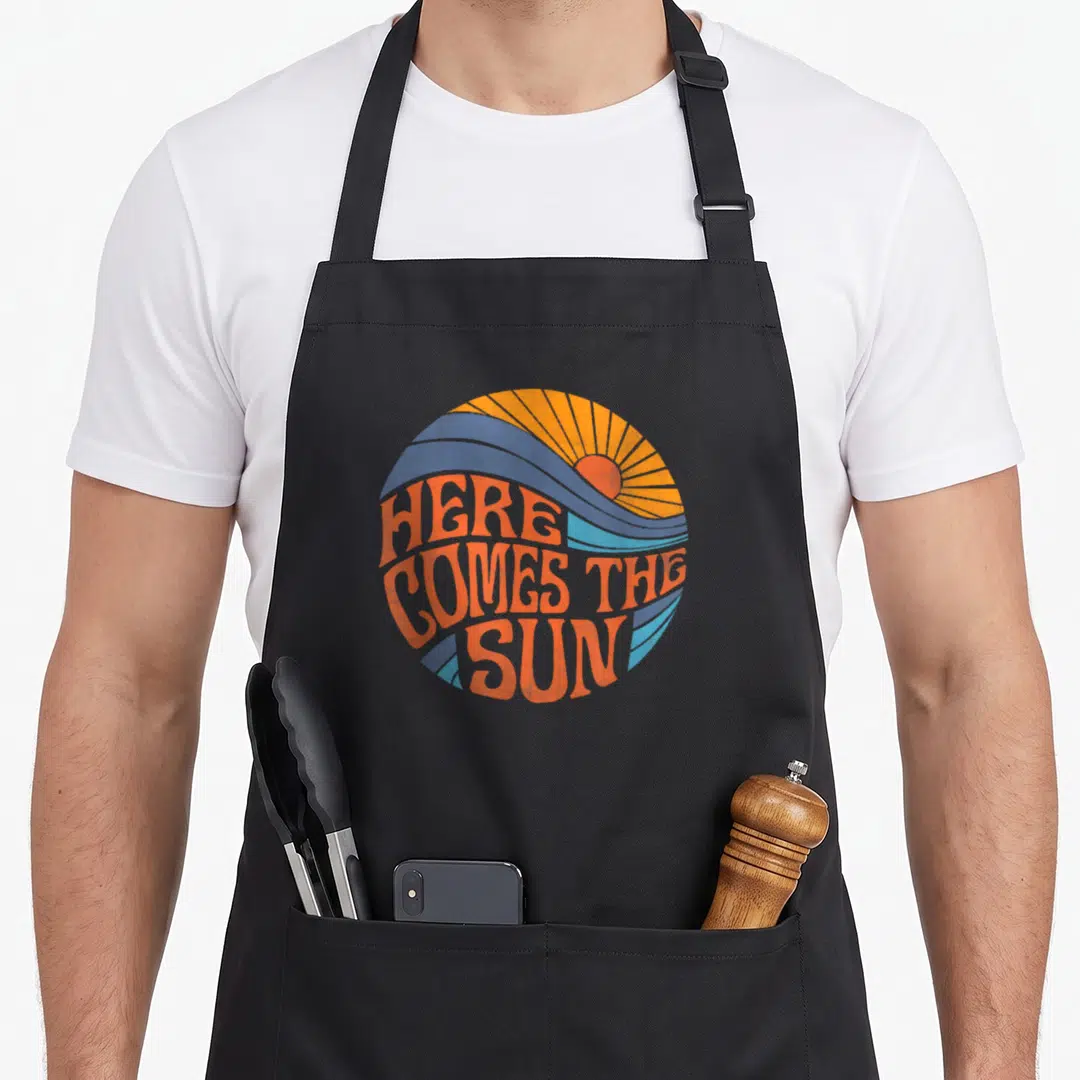 Sun come and get it apron - Retro circular print with orange sun, blue-grey waves, and bold orange text reading HERE — shown on a black cotton canvas apron against a white background