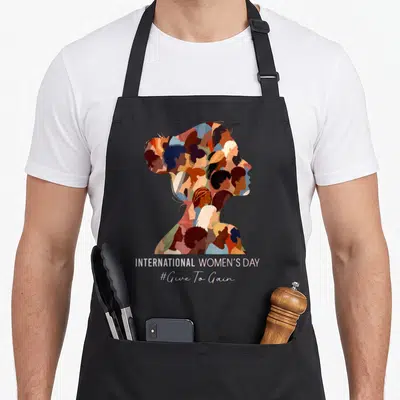International gains apron - Woman silhouette filled with smaller faces and stacked text INTERNATIONAL WOMEN'S DAY #Give — shown on a black cotton canvas apron against a white background