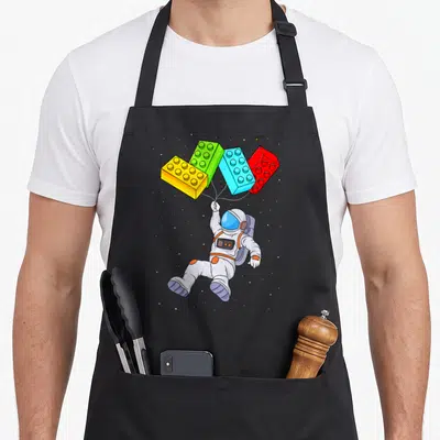 Space grill master apron - Cartoon astronaut floating below four toy bricks in white, yellow, green, blue, and red — shown on a black cotton canvas apron against a white background