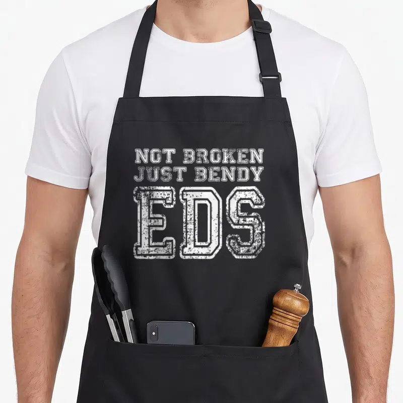 Broken eds awareness apron - White distressed text reading NOT BROKEN JUST BENDY EDS stacked vertically — shown on a black cotton canvas apron against a white background