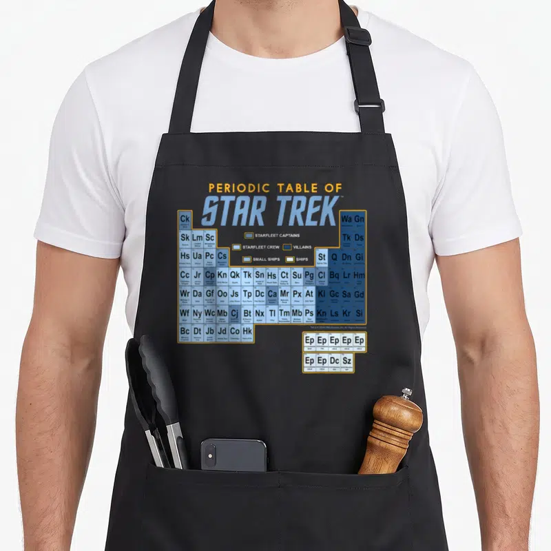 Original table apron - Periodic Table of Star Trek grid with character initials and ship names in yellow and blue cells — shown on a black cotton canvas apron against a white background