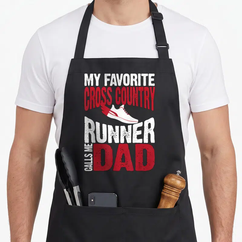 Call country apron - Running shoe graphic with text MY FAVORITE CROSS COUNTRY RUNNER CALLS ME DAD in white and red — shown on a black cotton canvas apron against a white background