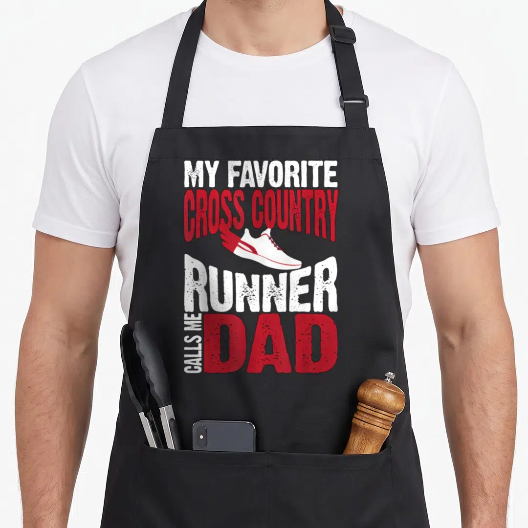 Call country apron - Running shoe graphic with text MY FAVORITE CROSS COUNTRY RUNNER CALLS ME DAD in white and red — shown on a black cotton canvas apron against a white background