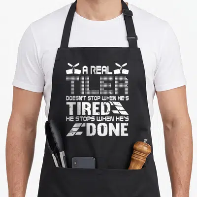 Setter tile apron - White trowels and grid-text reading A REAL TILER DOESN'T STOP WHEN HE'S TIRED — shown on a black cotton canvas apron against a white background