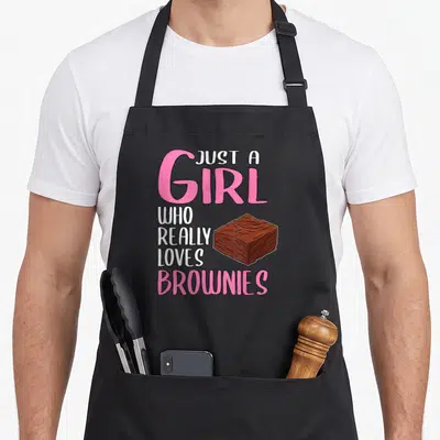 Brownie browny apron - Cartoon brownie next to stacked pink and white text reading Just A Girl Who Really Loves Brownies — shown on a black cotton canvas apron against a white background