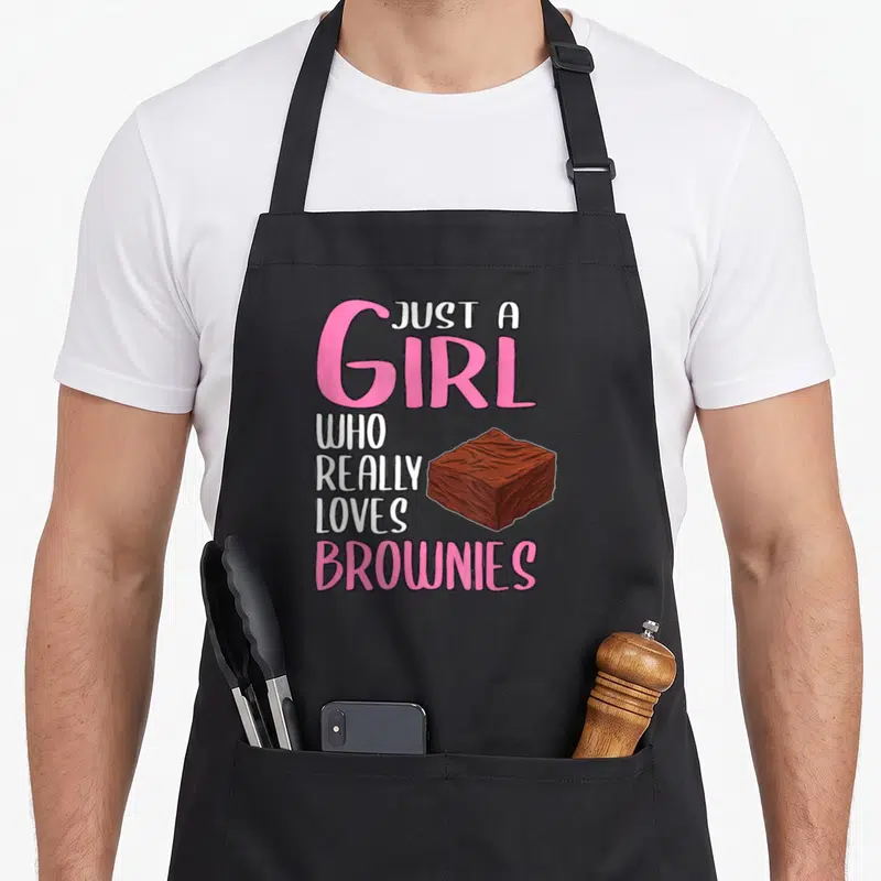 Brownie browny apron - Cartoon brownie next to stacked pink and white text reading Just A Girl Who Really Loves Brownies — shown on a black cotton canvas apron against a white background