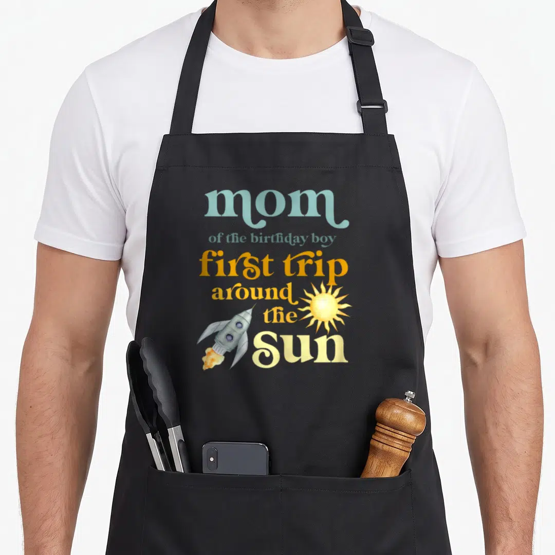 Sun birthday apron - Rocket and sun graphic with mom of birthday boy first trip around the Sun text — shown on a black cotton canvas apron against a white background