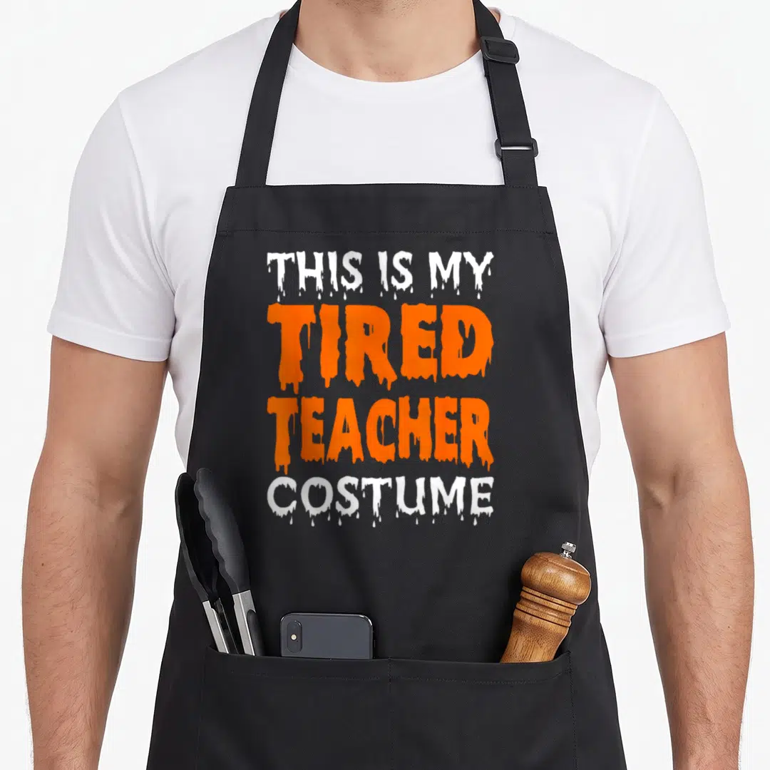 Tired quick apron - Dripping white and orange text reading THIS IS MY TIRED TEACHER COSTUME — shown on a black cotton canvas apron against a white background