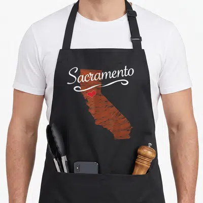Funny california sacramento apron - Reddish-brown California map with white Sacramento script and red heart marker — shown on a black cotton canvas apron against a white background
