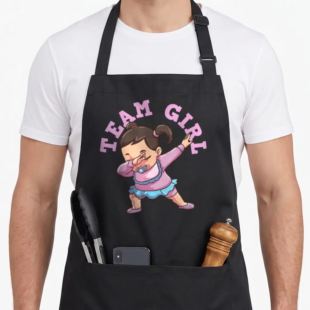 Cute gender reveal apron - Cartoon baby girl dabbing with TEAM GIRL text in pink and blue — shown on a black cotton canvas apron against a white background