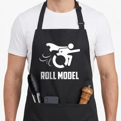 Roll wheelchair apron - White superhero silhouette in wheelchair with speed lines above ROLL MODEL text — shown on a black cotton canvas apron against a white background