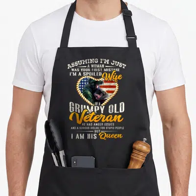 Spoiled grumpy wife apron - Distressed text surrounds rope heart with soldier child photo on USA flag in white yellow — shown on a black cotton canvas apron against a white background