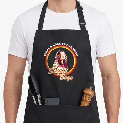 Funny max stupid aprons - Woman portrait inside red and yellow concentric circles with white arched text above — shown on a black cotton canvas apron against a white background
