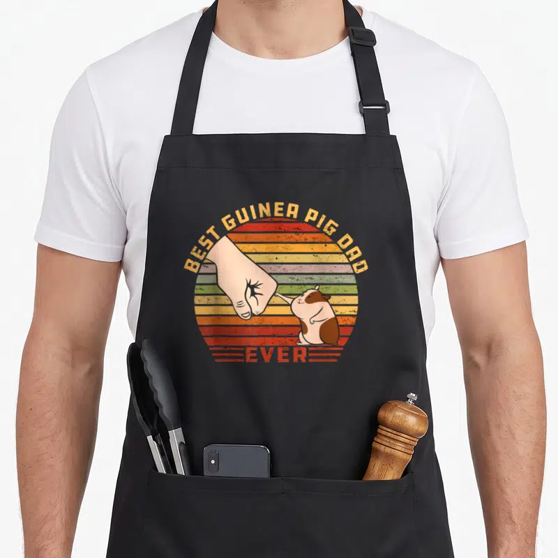 Father guinea pigs apron - Best Guinea Pig Dad Ever text with fist bumping cartoon cavy on striped sunset background — shown on a black cotton canvas apron against a white background
