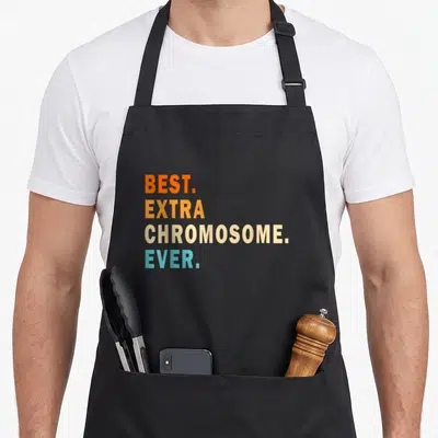 Awareness an extra apron - Stacked text BEST EXTRA CHROMOSOME EVER in orange yellow beige and blue — shown on a black cotton canvas apron against a white background