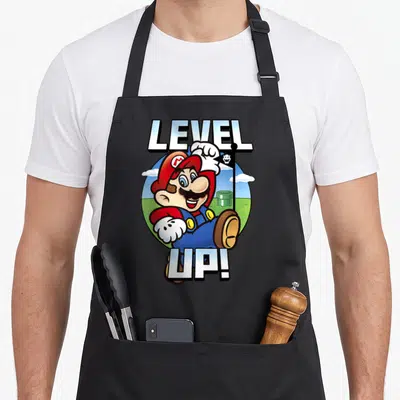 Level novelty apron - Mario holding a skull flag inside a circle with LEVEL UP! text above and below — shown on a black cotton canvas apron against a white background