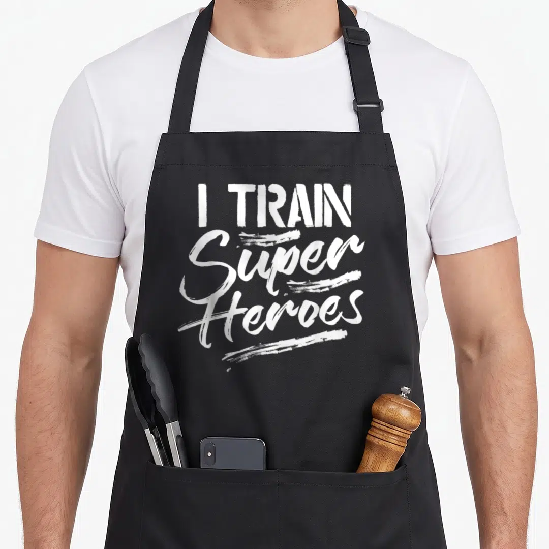 Train personalized teacher apron - White stacked typography reading I Train Super Heroes with brush strokes on black — shown on a black cotton canvas apron against a white background