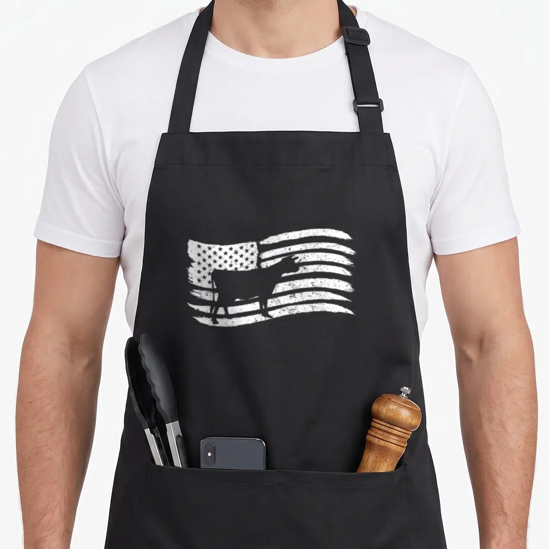Flag cow apron - White cow silhouette superimposed over a distressed American flag pattern — shown on a black cotton canvas apron against a white background