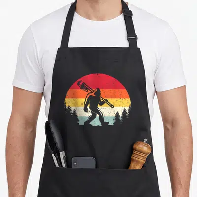 Plumber bigfoot apron - Grey Bigfoot silhouette with wrench on retro sunset background with pine trees — shown on a black cotton canvas apron against a white background