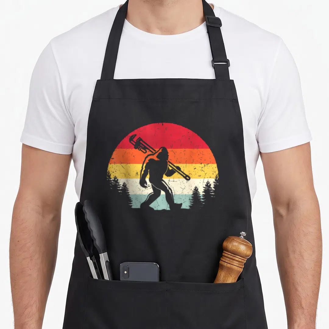 Plumber bigfoot apron - Grey Bigfoot silhouette with wrench on retro sunset background with pine trees — shown on a black cotton canvas apron against a white background