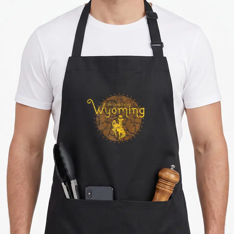 University floral apron - Yellow-orange circle with cowboy silhouette and text UNIVERSITY OF Wyoming — shown on a black cotton canvas apron against a white background