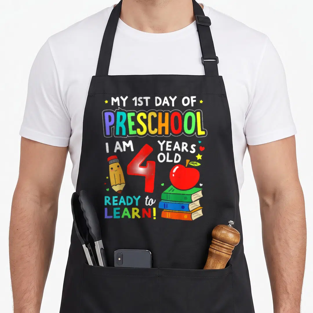 1st prek preschool apron - Cartoon pencil, red number 4, apple on books, and stacked text reading My 1st Day — shown on a black cotton canvas apron against a white background