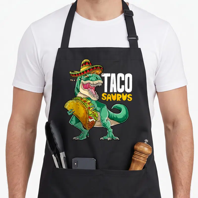 Taco dinosaur apron - Green T-Rex in sombrero holding taco with TACO SAURUS text — shown on a black cotton canvas apron against a white background